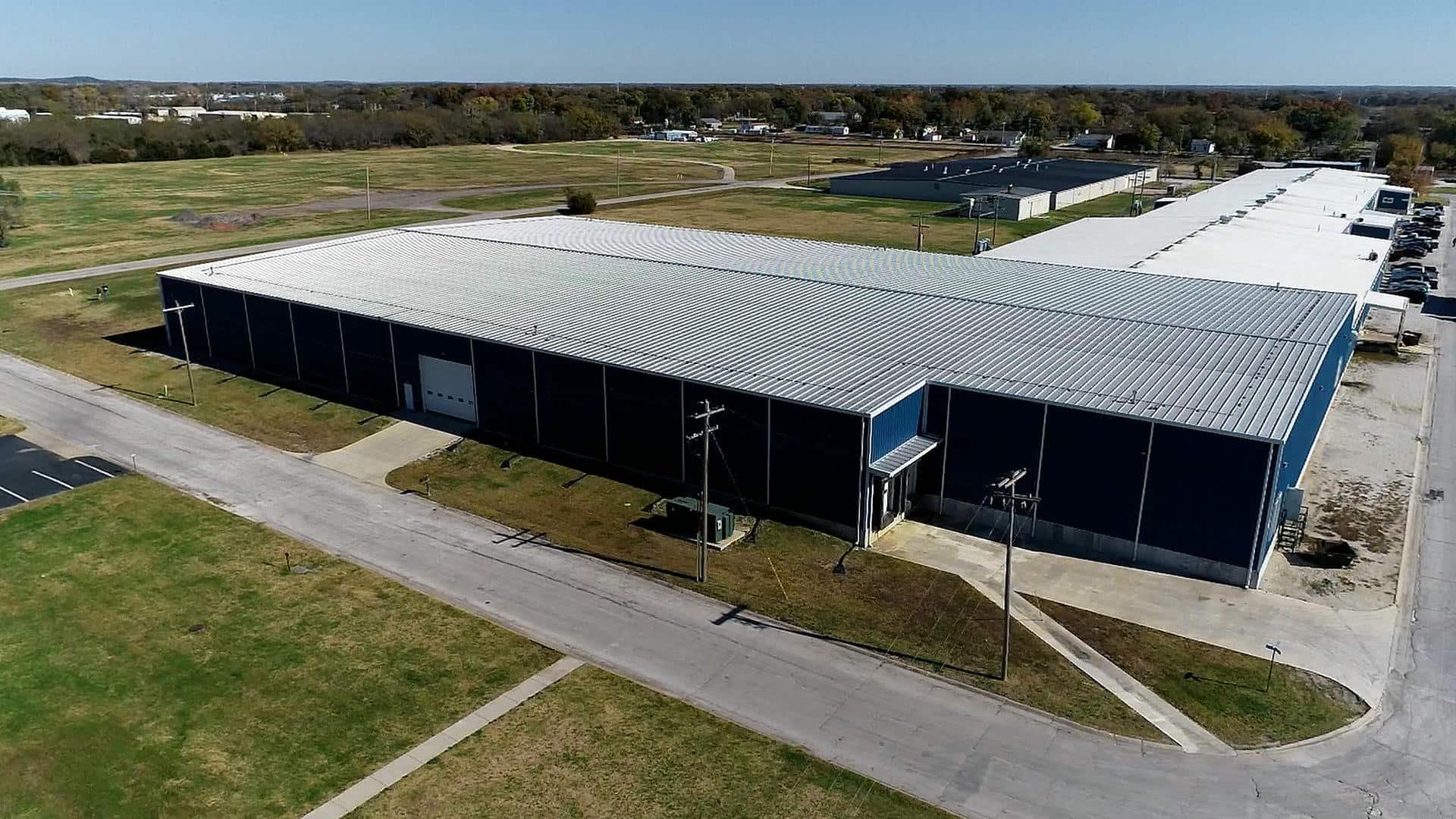 Aerial view of a large, blue industrial warehouse surrounded by empty roads and grassy fields under a clear blue sky, conveying a sense of isolation.