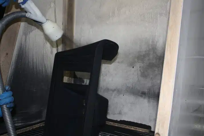 A person wearing blue gloves is using a spray gun to coat a black object in a booth. Gray walls show signs of previous spray activity.