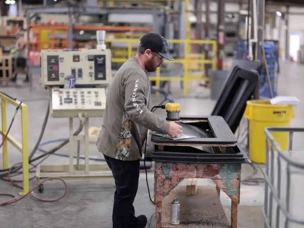 A man in a workshop uses a power tool on a metal sheet atop a makeshift table. Machinery and equipment are visible in the busy industrial setting.