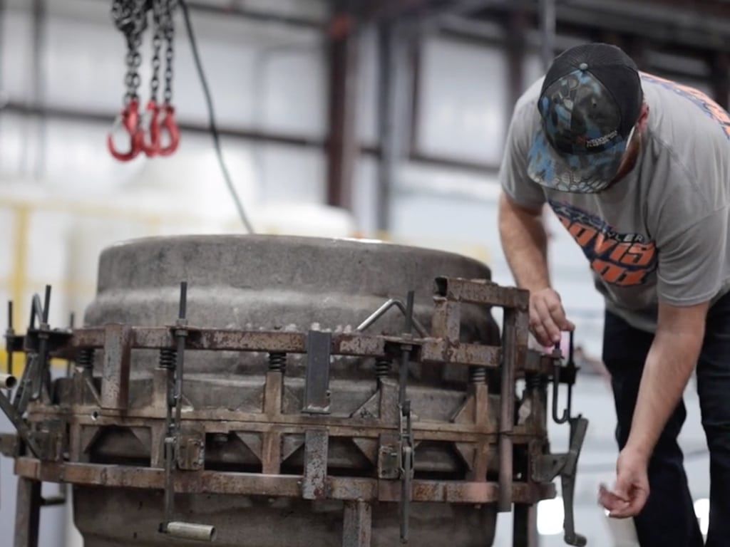 A person in a cap adjusts a large industrial mold with clamps in a workshop. Hanging chains are visible, conveying focus and precision work.