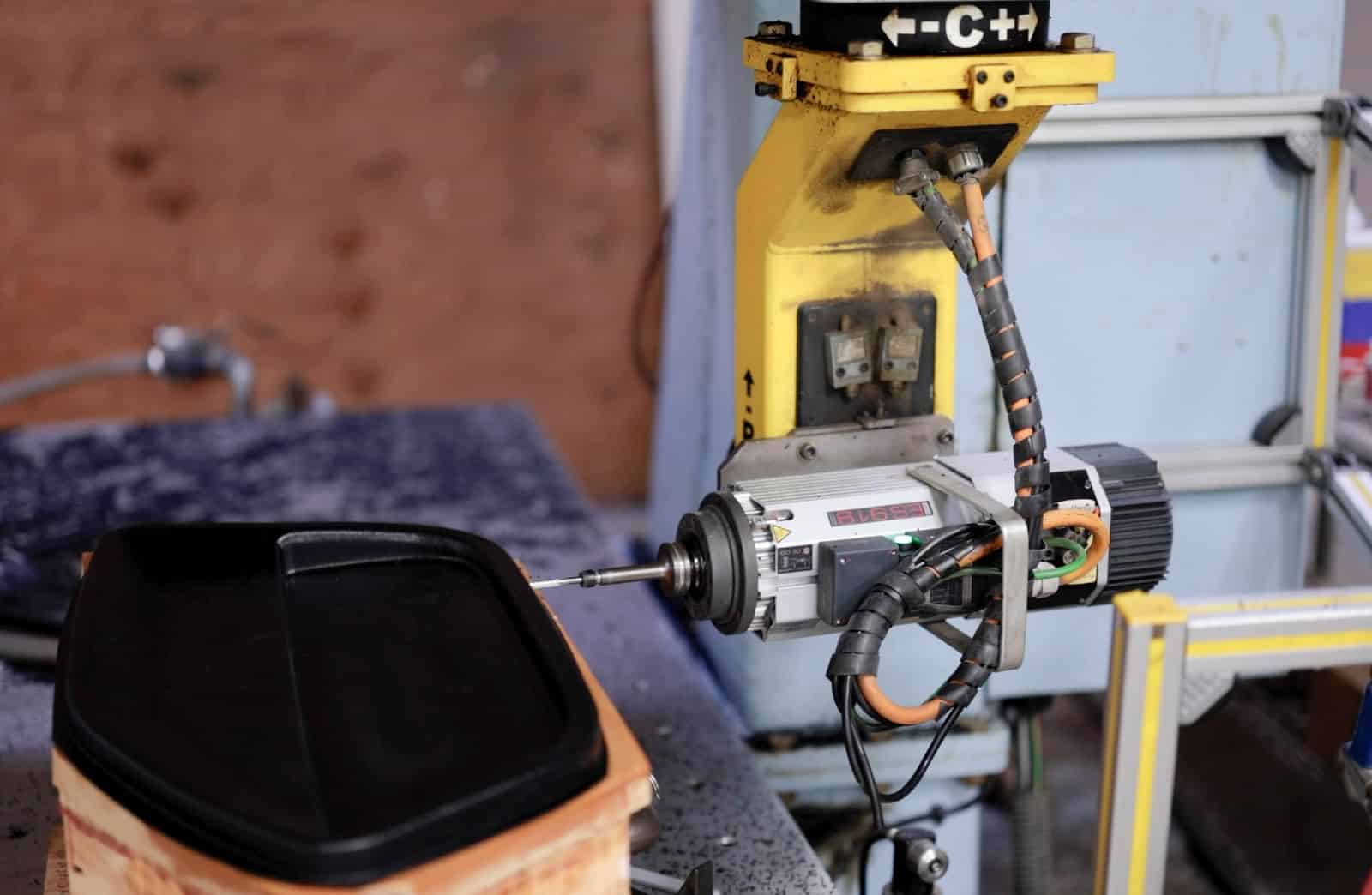 Robotic arm with a drill attachment poised above a black plastic mold on a workbench, suggesting precision manufacturing in an industrial setting.