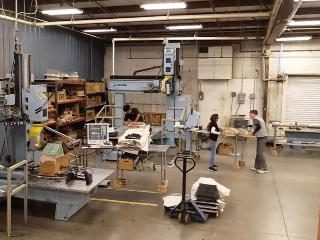 Factory floor with three people working at a CNC machine. Shelves are stacked with tools and boxes. The scene conveys a busy and industrious atmosphere.