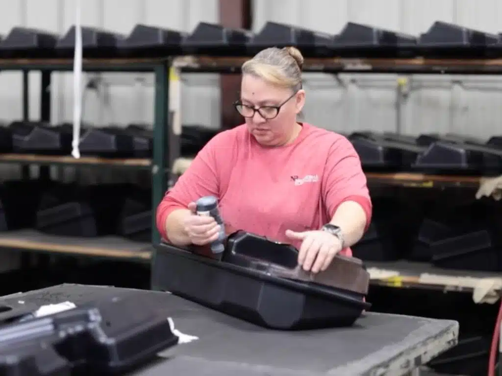 A person in a red shirt works on a black plastic part at a factory table, using a tool. Shelves in the background hold similar parts, suggesting a focused industrial setting.